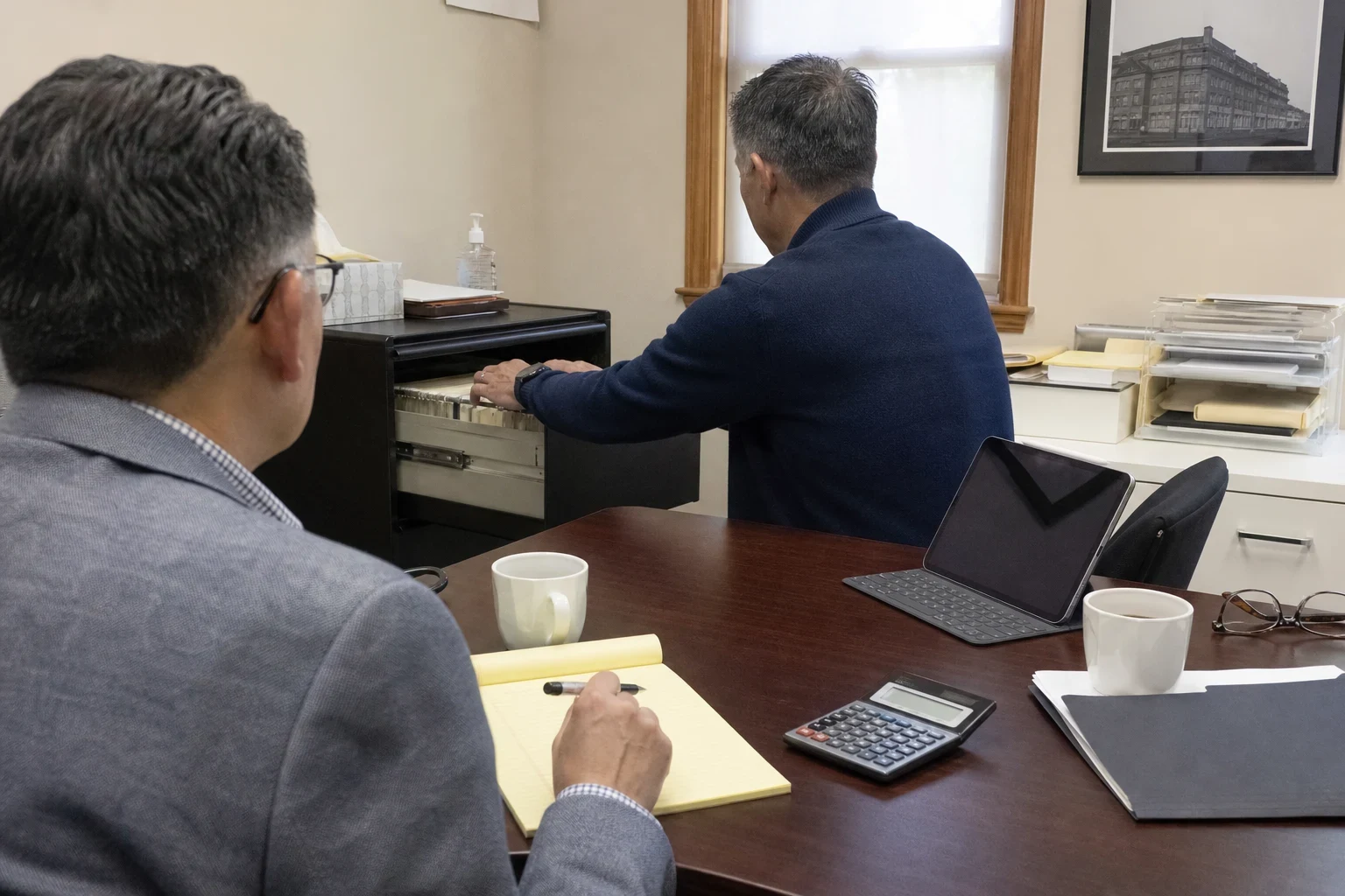 An agent with a yellow notepad and pen waits at a desk with a calculator and coffee while a man pulls a file from a filing cabinet drawer in a small office.