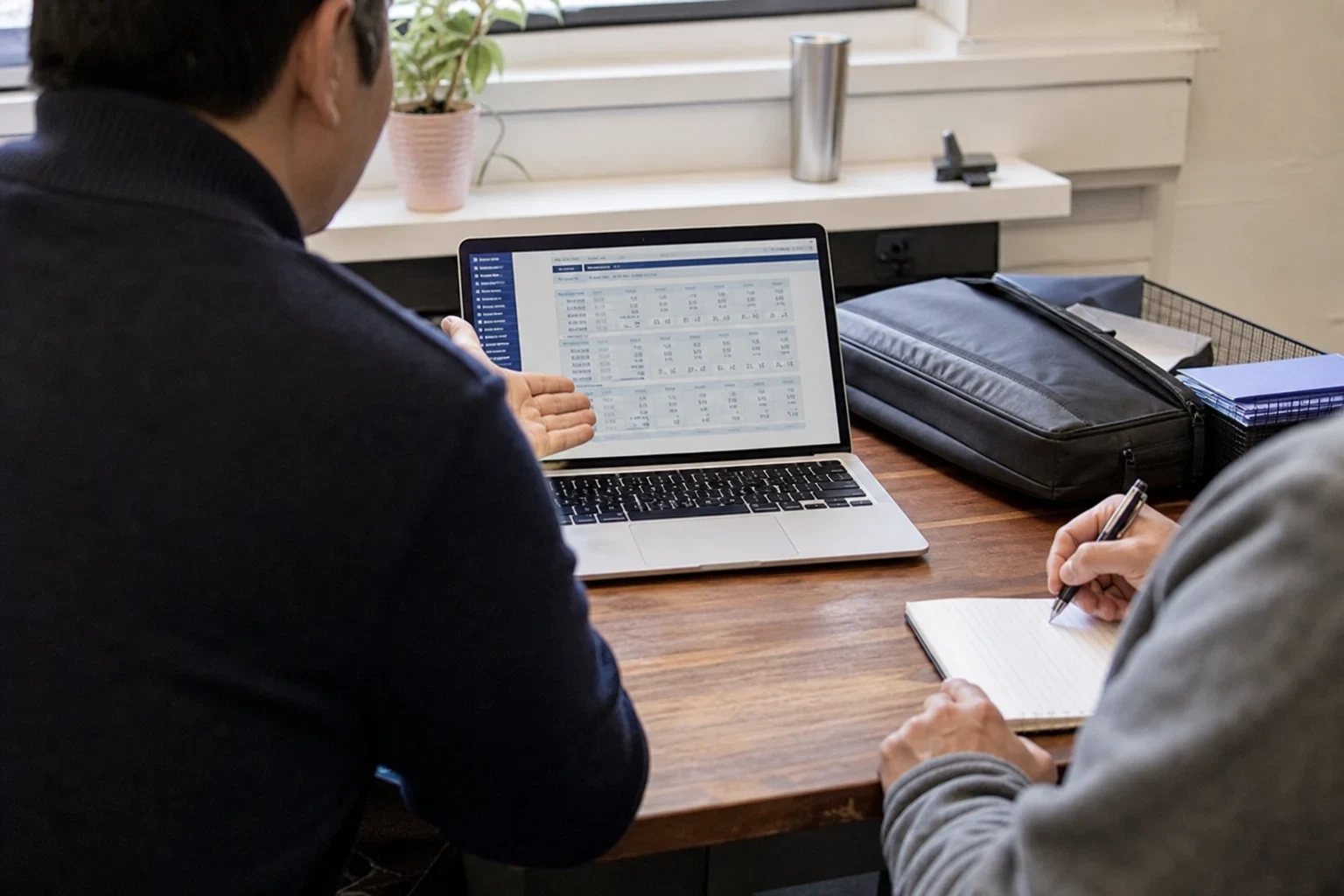 Over-the-shoulder view of a man gesturing at a laptop showing a table of figures on a wooden desk while another person takes notes in a notebook by a window