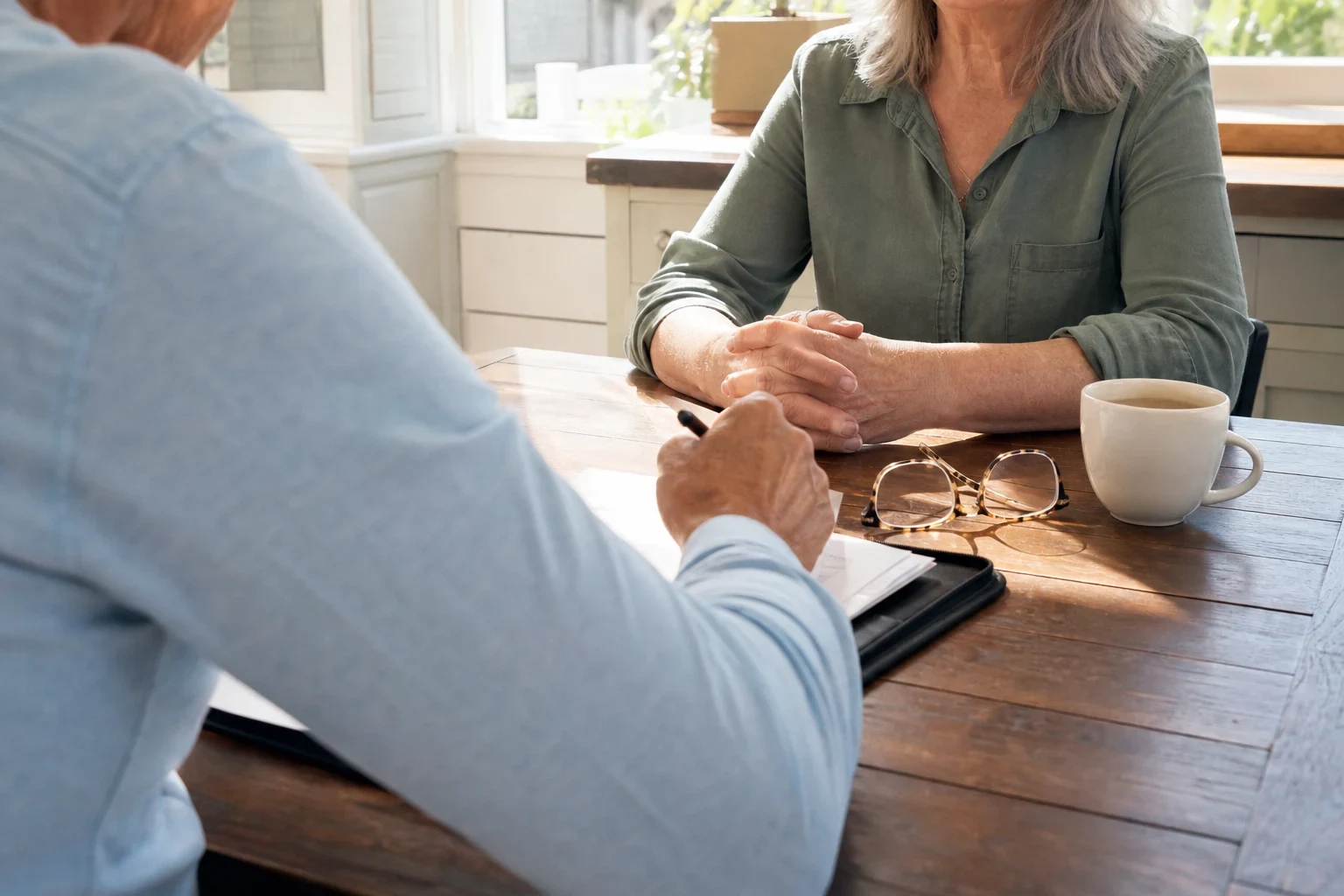 Agent in a light blue shirt takes notes in a padfolio across a wooden kitchen table from a gray-haired woman with clasped hands, eyeglasses and a coffee cup