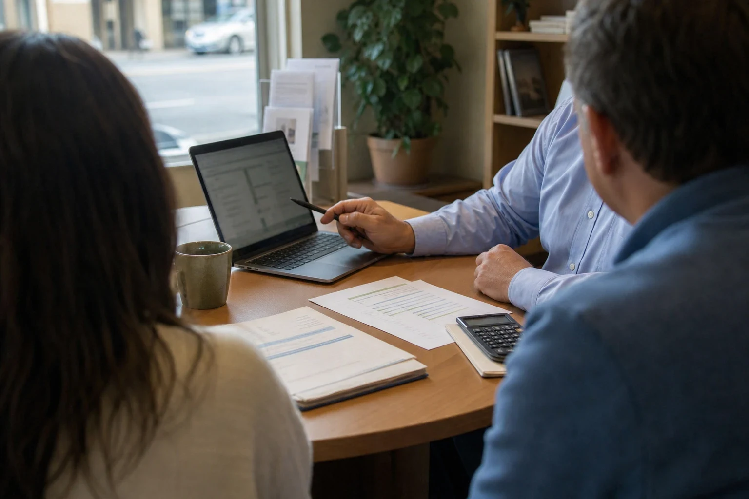 Over-the-shoulder view of a couple at a round office table as an agent in a light blue shirt points a pen at a laptop, with papers, a calculator and a plant by the window